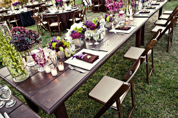 Table setting at outdoor reception - Purple, dark pink, and light green centerpieces, ivory candles, and maroon napkins with lavender decor and ivory floral accents on wooden table -  photo by Orange County based wedding photographers Mark Brooke